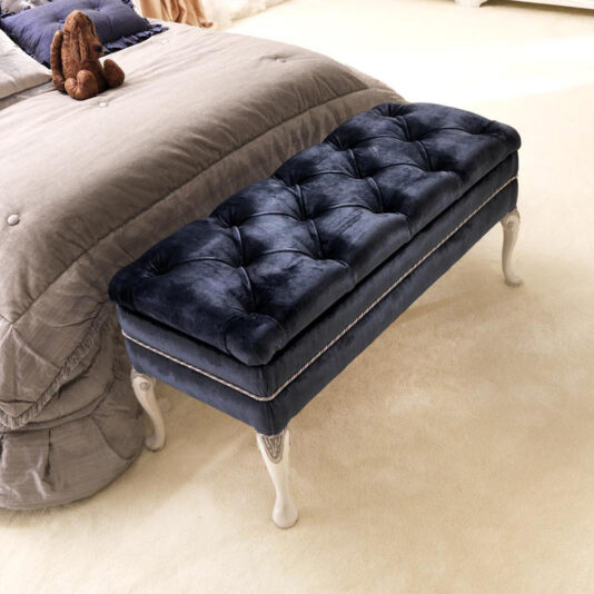 An Italian Button Upholstered Storage Bench in tufted navy-blue velvet with white legs is placed at the foot of a neatly made bed adorned with gray bedding. Near the pillows on the bed lies a small brown stuffed toy, and the room features a light-colored carpet.