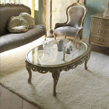 An elegant living room with classic furniture, featuring an Italian Reproduction Oval Glass Coffee Table with intricately carved legs, a vintage sofa with plush pillows, and an ornate armchair. A decorative chest of drawers stands beside the armchair, all atop a plush white rug.