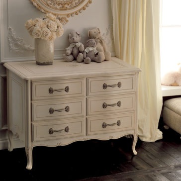 Small Italian Ivory Chest of Drawers An ornate vintage dresser with six drawers and floral handles stands below an elegant, decorative mirror. A vase of white roses and two plush toys rest on the Small Italian Ivory Chest of Drawers. Yellow curtains adorn the window to the right, casting soft light into the room.