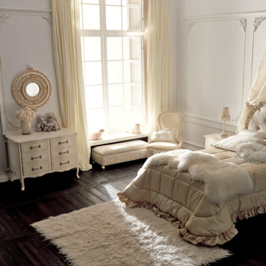 Small Italian Ivory Chest of Drawers Elegant bedroom with a large, sunlit window framed by cream-colored curtains. A plush bed adorned with a white fur throw sits alongside an ornate dresser featuring a circular mirror and a flower vase. The cozy arrangement is complemented by an armchair, matching pouf, and the highlight—a Small Italian Ivory Chest of Drawers—placed on a soft white rug atop dark wood flooring to complete the room's decor.