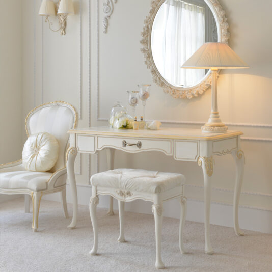 A vintage vanity setup features the Classic Ivory and Gold Italian Dressing Table with its matching stool, a round carved mirror, and a classic table lamp. An elegant upholstered chair is placed nearby. The scene is set in a room with light decor and soft furnishings.
