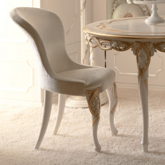 A bright, light-filled room with plush carpeting features a Designer Upholstered Italian Chair, showcasing its luxurious and elegant design with a curved back and gold trim. Nearby, an ornate round table adorned with intricate carvings and gold accents adds to the sophistication of the space.