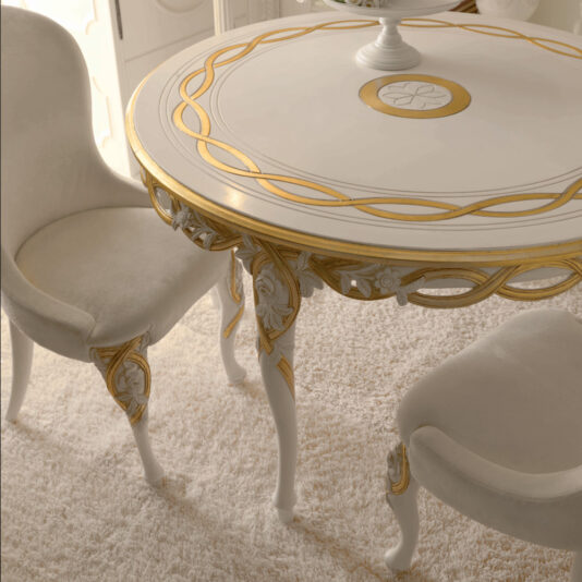 Designer Upholstered Italian Chair A close-up of an elegant round table with ornate gold detailing on a white base. Two Designer Upholstered Italian Chairs, featuring a matching gold and white design, are positioned next to the table, all set on a light-colored plush carpet.