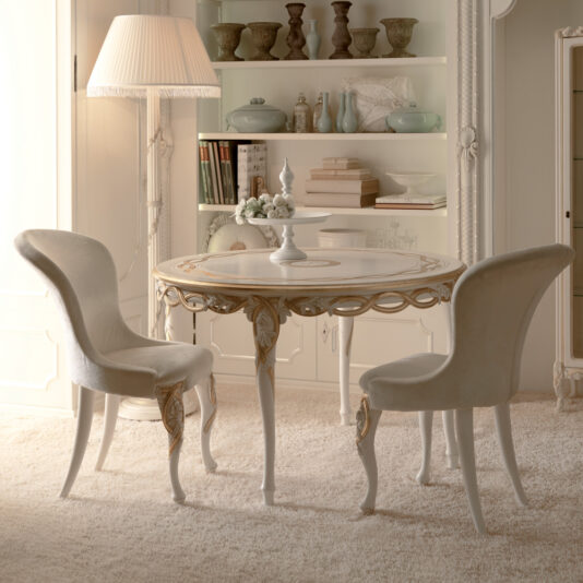 Designer Upholstered Italian Chair A luxurious, elegantly decorated room with a round table and two Designer Upholstered Italian Chairs. The table features intricate designs and a centerpiece of flowers. Behind the table, a white bookcase filled with books, decorative ceramics, and delicate artifacts adds to the sophisticated atmosphere.