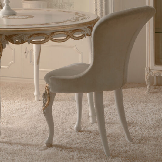 Designer Upholstered Italian Chair An elegant, classic dining area showcasing a round table with intricate gold detailing and a Designer Upholstered Italian Chair featuring distinctive curved legs. The scene is complemented by a beige plush carpet and luxurious off-white surroundings adorned with ornate furnishings.