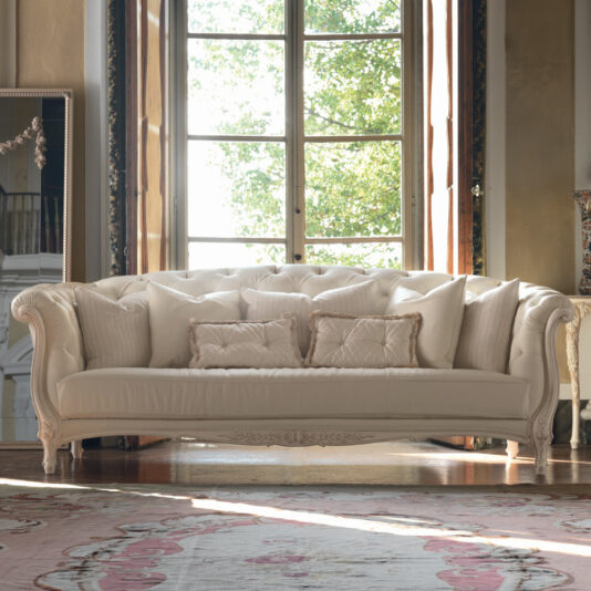 A luxurious Reproduction Baroque Italian Leather Large 3 Seater Sofa adorned with multiple matching pillows sits in front of a large window with wooden frames, revealing lush green trees outside. The elegant room features ornate details and a richly patterned rug on the wooden floor.
