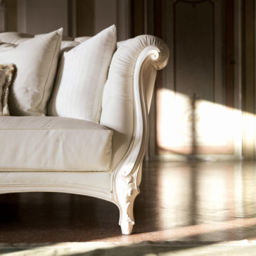 Reproduction Baroque Italian Leather Large 3 Seater Sofa A close-up of the arm of a luxurious Reproduction Baroque Italian Leather Large 3 Seater Sofa reveals ornate carved wood details. The sofa is adorned with white, plush cushions, while sunlight streams through the room, casting a warm glow on the polished wooden floor and elegant furnishings.