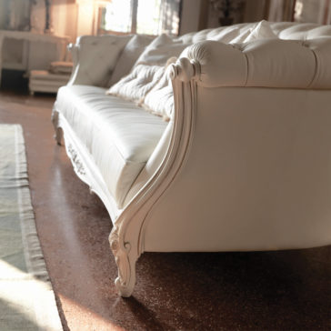 Reproduction Baroque Italian Leather Large 3 Seater Sofa A beautifully upholstered Reproduction Baroque Italian Leather Large 3 Seater Sofa with a tufted backrest and ornate wooden legs sits in a sunlit room. The design is highlighted as the light catches the elegant white leather. The polished brown floor and a light-colored rug partially visible at the bottom left complete the scene.