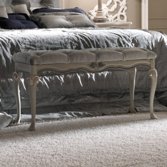 A luxurious bedroom showcases the Designer Italian Button Upholstered Bedroom Bench with ornate wooden legs, positioned at the foot of a plush, silvery-gray bed. Both the bench and the nearby nightstand feature elaborate, carved details that enhance the room's elegant decor.