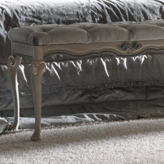 An elegant Designer Italian Button Upholstered Bedroom Bench with ornate legs stands on a plush, light-colored carpet. The tufted bedroom bench is upholstered in a soft, velvet-like fabric, and the backdrop showcases a luxurious, silky draped fabric, enhancing the refined atmosphere.