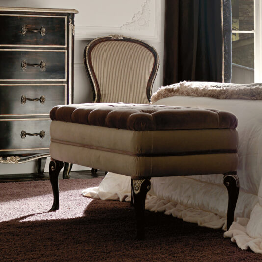 An elegant bedroom features a Designer Italian Button Upholstered Storage Bench with dark wooden legs at the foot of a bed adorned with a white comforter. Against the wall, there is a classic wooden chest of drawers with ornate handles and a vintage upholstered chair. The room is softly illuminated by natural light.