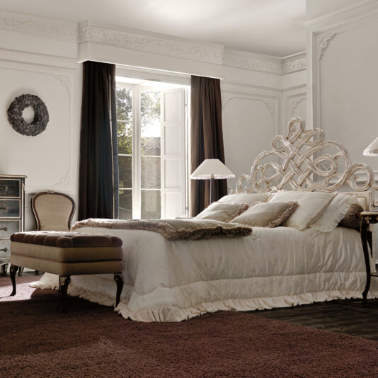 Elegant bedroom with a large ornate bed, a Designer Italian Button Upholstered Storage Bench at the foot, and two bedside lamps. Tall windows with dark curtains let in natural light. The room features white walls, crown molding, a rug, a chest of drawers, and a decorative wreath.
