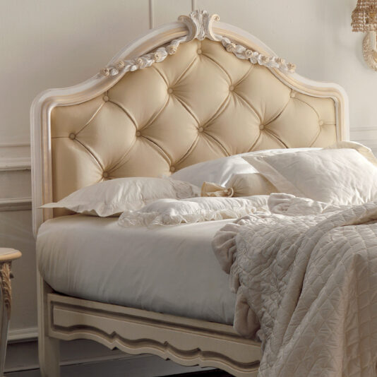 The Italian Rose Detail Luxury Single Bed features an elegant design with a plush, cream-colored, tufted headboard. It is neatly made with white and beige bedding, including a quilted comforter and several pillows. The ornate carvings on the frame showcase exquisite Italian rose details, creating a cozy and luxurious setting.