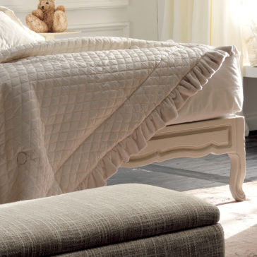 Italian Rose Detail Luxury Single Bed The Italian Rose Detail Luxury Single Bed takes center stage in this cozy bedroom scene, adorned with a neatly made quilted, cream-colored coverlet featuring delicate ruffled edges. A small teddy bear adds a charming touch atop the headboard. The room also includes an upholstered ottoman and is softly illuminated by natural light filtering through window curtains.