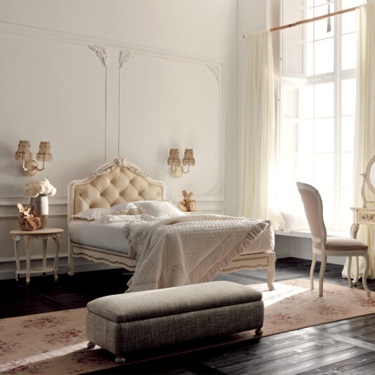 A bright, elegant bedroom showcases the Italian Rose Detail Luxury Single Bed with its plush, tufted headboard and neatly made cream-colored quilt adorned with delicate Italian rose details. The vintage-style decor includes a matching nightstand, a vanity with a mirror, and a cushioned bench. Large windows with sheer curtains let natural light flood the room.