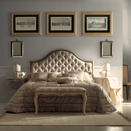 A luxurious bedroom features a tufted headboard and a plush bed adorned with cushions and a quilted bedspread. The Luxurious Designer Italian Bedroom Bench sits gracefully at the foot of the bed, flanked by two small tables with lamps. Above the bed are three framed pictures and two smaller mirrors, completing the elegant, muted decor.