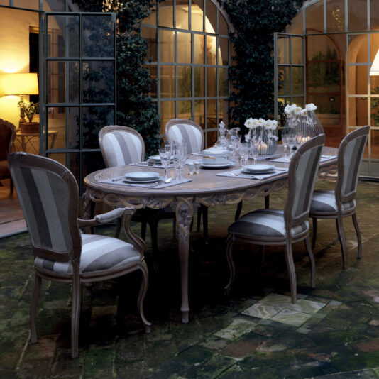 Elegant outdoor dining area featuring the Luxurious Italian 6 Seat Dining Table Set, with a long, ornately carved wooden table adorned with plates, glasses, and white flowers in a vase. Six upholstered chairs with striped fabric surround the table. Large windows with arched frames and ivy-covered walls complete the scene.