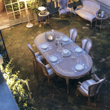 An elegant outdoor dining set on a brick patio features the Luxurious Italian 6 Seat Dining Table Set with intricate designs, six cushioned chairs, and place settings for six. A plush sofa and armchair set are in the background beside potted plants, creating a cozy and inviting atmosphere.