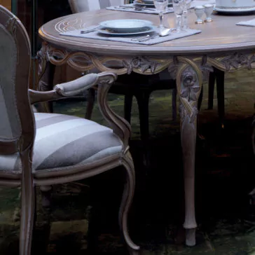 The Luxurious Italian 6 Seat Dining Table Set showcases fine china and crystal glassware, with intricate carvings adorning both the wooden table and chairs. The cushioned seats are upholstered in light fabric, while the table is elegantly accented by a detailed floral design.