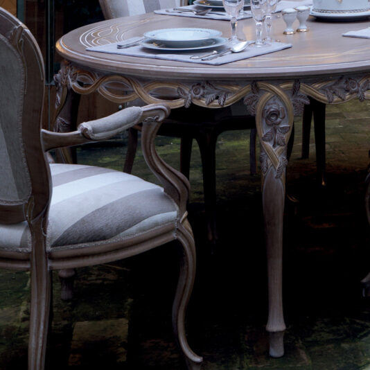 The Luxurious Italian 6 Seat Dining Table Set showcases fine china and crystal glassware, with intricate carvings adorning both the wooden table and chairs. The cushioned seats are upholstered in light fabric, while the table is elegantly accented by a detailed floral design.