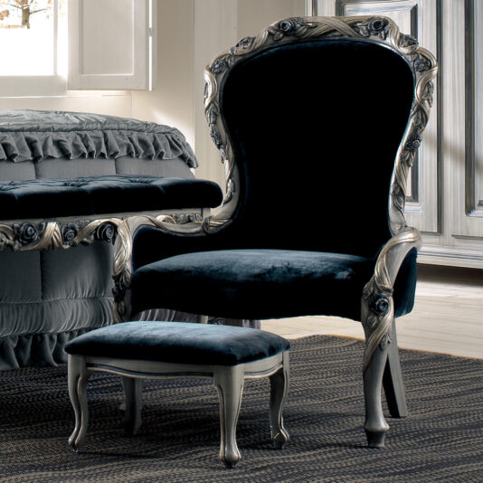 An ornate, dark velvet Luxury Italian Ladies Bedroom Chair with a matching footstool sits on a patterned rug in an elegantly decorated room. The chair and footstool feature intricately carved wooden frames. A window with partially opened shutters and a bed with a ruffled coverlet are visible nearby.