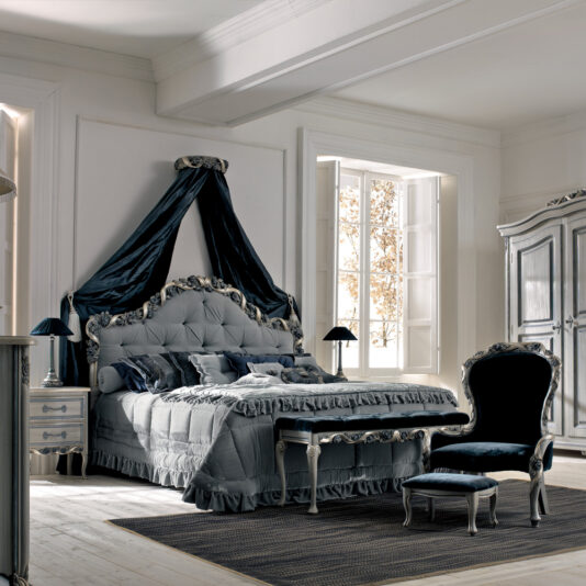 A lavishly decorated bedroom features a four-poster bed draped in dark blue fabric and adorned with an ornate headboard. It includes a matching Luxury Italian Ladies Bedroom Chair with footstool, positioned beside an elegant white wardrobe. Large windows provide ample natural light.