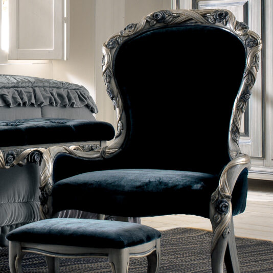 A luxurious setting features the opulent Luxury Italian Ladies Bedroom Chair, characterized by its dark upholstery and intricate wood carvings. In front of it stands a matching ottoman. The background showcases a ruffled bedspread, complemented by elegant, classic décor elements.