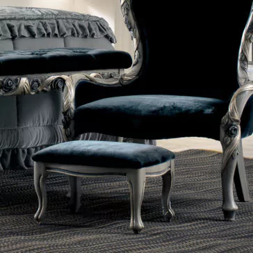 Luxury Italian Ladies Foot Stool An elegant dark blue velvet chair with an intricate silver frame paired with a Luxury Italian Ladies Foot Stool complements the classic room featuring a ruffled bedspread. The furniture rests on a textured rug, adding a touch of luxury to the space.