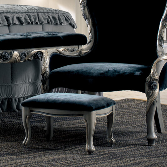 An elegant dark blue velvet chair with an intricate silver frame paired with a Luxury Italian Ladies Foot Stool complements the classic room featuring a ruffled bedspread. The furniture rests on a textured rug, adding a touch of luxury to the space.