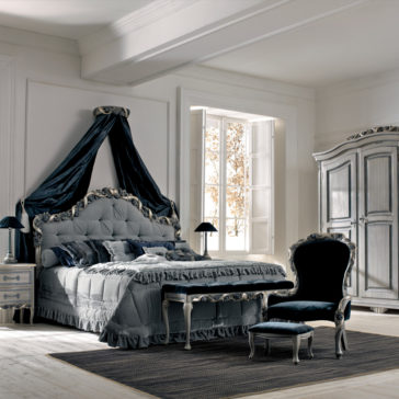 Luxury Italian Ladies Foot Stool An elegant bedroom with a plush, tufted bed adorned with a draped canopy. The room features a large window letting in soft light, an ornate wardrobe, a matching nightstand, and a cushioned chair with an ottoman. A Luxury Italian Ladies Foot Stool enhances the predominantly grey and blue decor with an extra touch of sophistication.