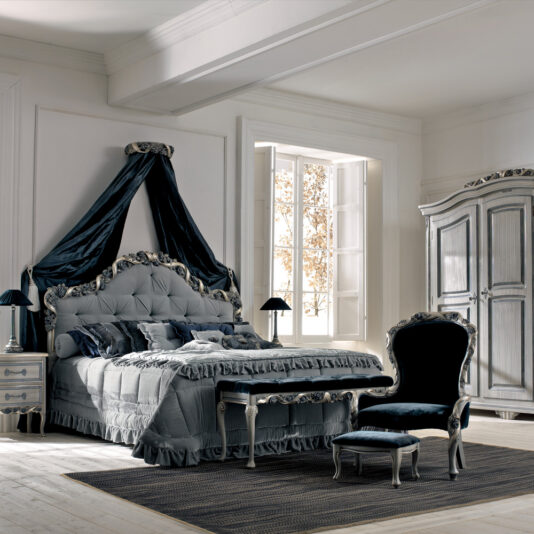 An elegant bedroom with a plush, tufted bed adorned with a draped canopy. The room features a large window letting in soft light, an ornate wardrobe, a matching nightstand, and a cushioned chair with an ottoman. A Luxury Italian Ladies Foot Stool enhances the predominantly grey and blue decor with an extra touch of sophistication.