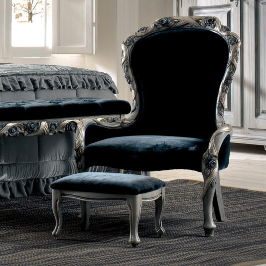 A luxurious Italian velvet armchair with ornate silver detailing is paired with a Luxury Italian Ladies Foot Stool in matching style. The set is placed on a dark woven rug in an elegant room. A bed with a ruffled skirt and a wardrobe in similar ornate style are visible in the background.