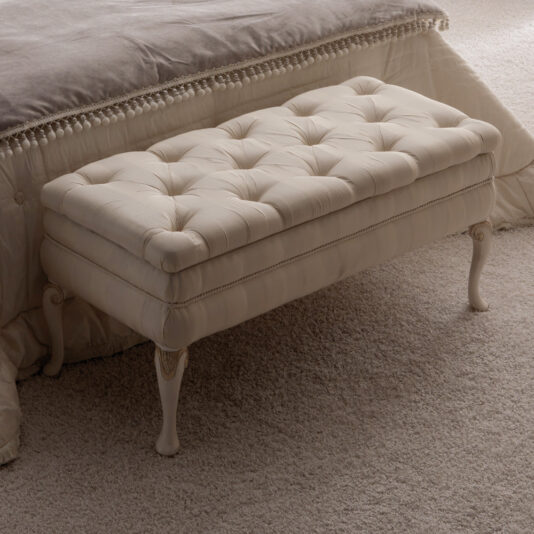 A Reproduction Designer Italian Button Upholstered Storage Bench with cream-colored tufted details and carved wooden legs is placed at the foot of a bed. The bed features a beige blanket with fringe detailing, and the floor is carpeted in a light shade. The setting appears to be a cozy and elegant bedroom.