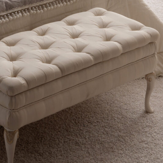 Reproduction Designer Italian Button Upholstered Storage Bench An elegant, cream-colored Reproduction Designer Italian Button Upholstered Storage Bench with decorative wooden legs stands on a plush, beige carpet. This exquisitely crafted bench features deep button details on the cushioned top and offers convenient storage, perfectly positioned near a white, textured bedspread.