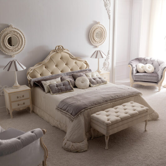 Reproduction Designer Italian Button Upholstered Storage Bench A luxurious bedroom in neutral tones features a tufted bed with plush pillows and throws, flanked by two ornate nightstands with matching lamps. Elegant armchairs and the Reproduction Designer Italian Button Upholstered Storage Bench add comfort, while round framed mirrors adorn the wall above the nightstands.