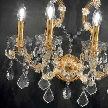 A close-up of an ornate chandelier with three lit candles. The fixture, similar to the 4 Light Venetian Wall Light, showcases gold accents and numerous dangling crystal prisms, creating a sparkling effect against a black background.
