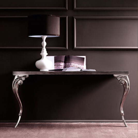 A Designer Italian Console Hall Table with ornate metallic legs graces a dark paneled wall, adorned with a white sculptural lamp featuring a black shade and an open magazine. The plush carpet adds to the room's sophisticated ambiance.