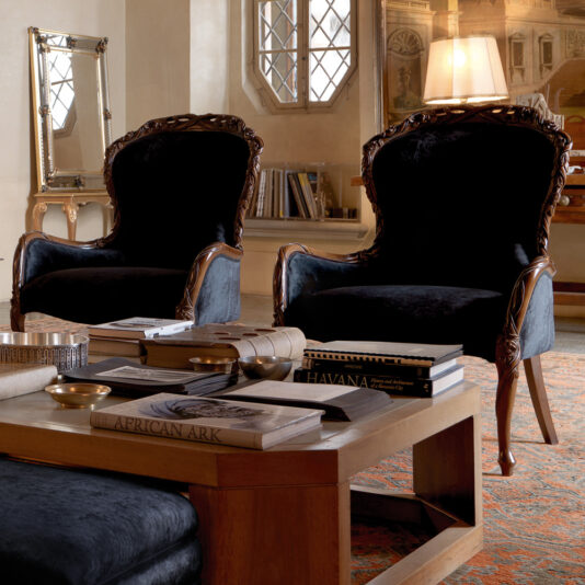 Two Classic Italian Rose and Ribbon Walnut Armchairs with dark velvet upholstery are placed in an elegant, traditional living room. A wooden coffee table in front of the chairs holds books and decorative items. A large window, artwork, and a lamp in the background complete the sophisticated setting.