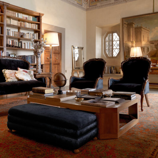 A luxurious living room with elegant décor, featuring a wooden coffee table, plush black seating, and a large bookshelf filled with books. The room is warmly lit by standing lamps and showcases the Classic Italian Rose And Ribbon Walnut Armchair, a large painting, and a decorative mirror on the walls.