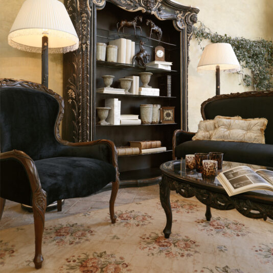 A cozy, elegant living room featuring a plush black sofa and a Classic Italian Rose And Ribbon Walnut Armchair, a dark wooden bookshelf adorned with decorative items and books, two tall lamps with white shades, a glass coffee table with books and mugs, and a floral-patterned rug.