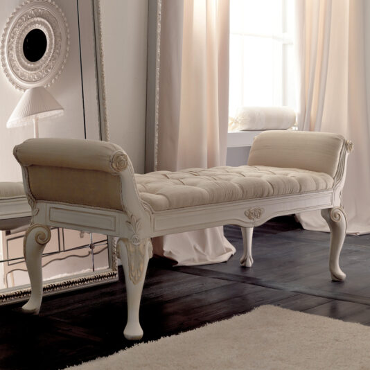 A stylish, cream-colored chaise lounge with tufted upholstery and intricately carved wooden legs is positioned next to a large wall mirror. The Designer Italian Button Upholstered Bedroom Bench complements the room's elegance. Sheer curtains allow natural light to fill the space, highlighting the dark wooden floor and light-colored rug.