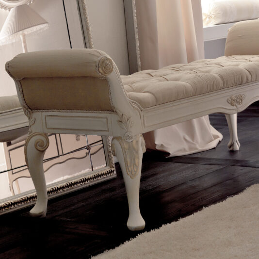 Designer Italian Button Upholstered Bedroom Bench A luxurious Designer Italian Button Upholstered Bedroom Bench with carved wooden legs is placed beside a large, decorative mirror on a dark hardwood floor. Cream-colored drapes hang in the background, and a plush rug partially covers the floor in front of the bench.