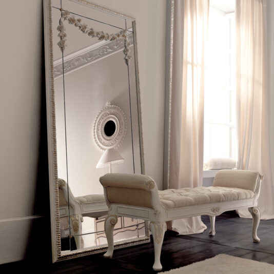 Designer Italian Button Upholstered Bedroom Bench An elegant room showcases a tall, ornate mirror leaning against the wall beside a large window adorned with flowing white curtains. Positioned in front of the mirror is a plush Designer Italian Button Upholstered Bedroom Bench, complete with intricate carvings on its legs and arms. The room is bathed in soft, natural light.