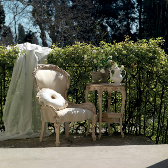A vintage-style outdoor scene featuring a Designer Italian Rose And Ribbon Armchair adorned with a white cushion. Beside the armchair, there's a small table displaying a potted plant and a white pitcher. A sheer white curtain elegantly flows over the hedge, with lush greenery and trees peeking through in the background.