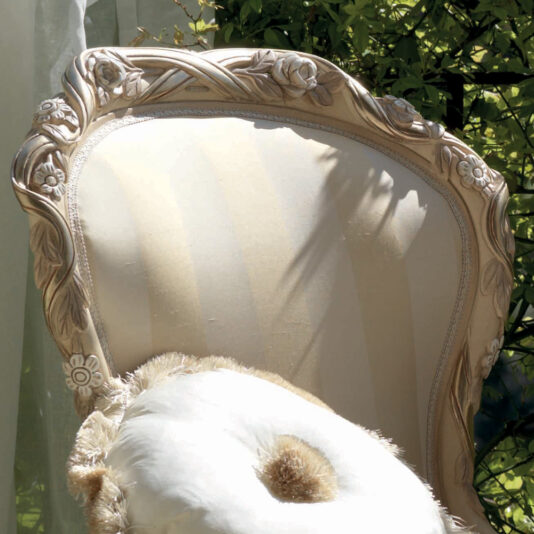 A beige upholstered Designer Italian Rose And Ribbon Armchair featuring ornate wooden carvings of flowers and leaves on its frame is placed outdoors. The chair is adorned with round, fluffy pillows and is partially shaded by sunlit greenery in the background.