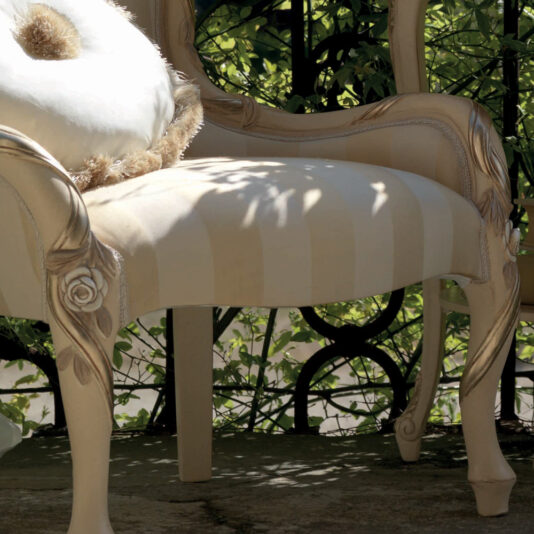 A close-up of a Designer Italian Rose And Ribbon Armchair featuring ornate carvings and a cream color. The floral details on the arms and legs highlight its craftsmanship. Resting on the seat is a round, white cushion adorned with shaggy brown fringe. In the background, a wrought iron railing intertwined with green vines adds to the elegant ambiance.