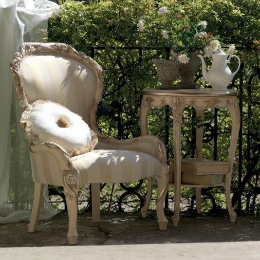A cozy outdoor seating area showcases a Designer Italian Rose And Ribbon Armchair adorned with a fluffy white pillow, positioned beside an elegant side table. The table is topped with a white teapot, a decorative vase, and a potted plant. A backdrop of green foliage enhances the serene setting.