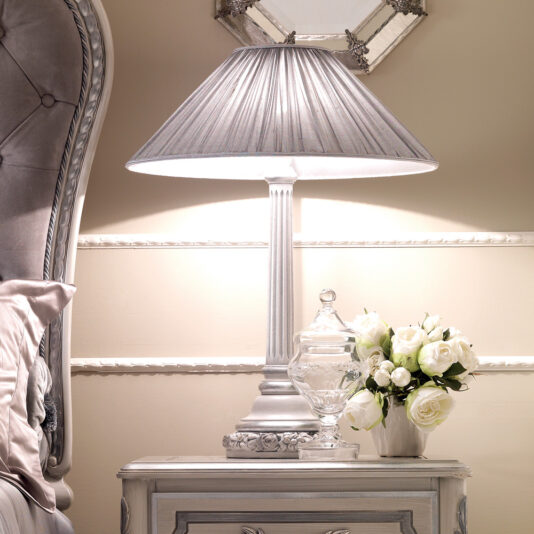 An elegant bedside table showcases the Designer Italian Rose Lamp with its pleated gray lampshade. A glass jar with a lid is placed next to a small white vase holding white roses. In the background, part of a decorative headboard and a framed art piece can be seen.