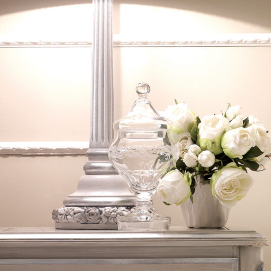 Designer Italian Rose Lamp A close-up of a decorative scene features a white table adorned with a tall, ornate Designer Italian Rose Lamp, a clear glass jar with a lid, and a white ceramic pot holding a bouquet of white roses and small white flowers. The background is softly lit and beige-colored.