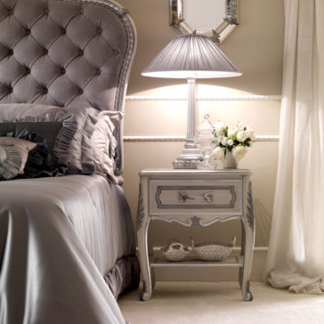 Designer Italian Rose Lamp In a luxurious bedroom, a tufted headboard bed is adorned with satin pillows and sheets. A vintage bedside table showcases the Designer Italian Rose Lamp, along with a small jar and vase of white roses. The room is elegantly decorated with beige walls and soft curtains.